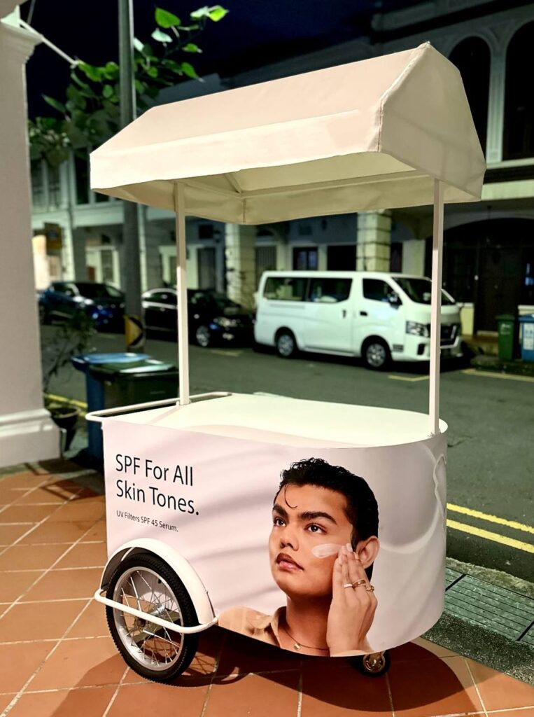 Off-white mobile pushcart with canopy roof, comes with large storage compartment. This cart is wrapped with artwork from The Ordinary skincare brand. Fully customisable from cart body wrapping, simple logo customisation to printing of logo on the cart roof. Our pushcart is available in both black and off white. Perfect for reception tables, product displays, coffee cart, flower bars, or as a centerpiece to any event.