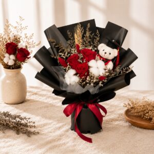 A graduation bouquet featuring preserved red roses, cotton blooms, dried florals, and a mini graduation bear! Bold and sophisticated, perfect for celebrating a milestone achievement.