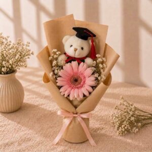 Single-stalk fresh pink gerbera, accompanied by fresh white limonium, wrapped in rustic brown kraft paper, topped with white graduation bear